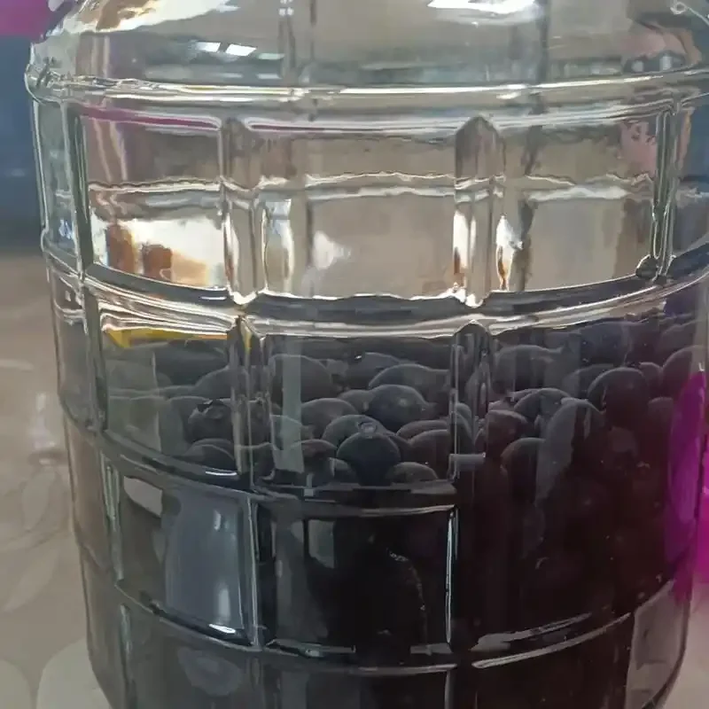 家庭自制蓝莓酒过程展示：透明玻璃罐中装满蓝莓、冰糖与酒液，旁边有新鲜蓝莓和工具_1