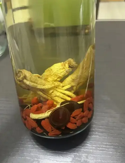 透明玻璃泡酒瓶内泡制的鹿茸人参枸杞酒，色泽金黄透亮，旁边散落着称量工具和药材，展示家庭泡酒的专业场景。_1