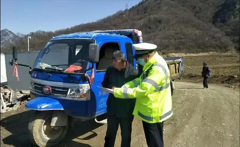 一位驾驶员站在一辆蓝色三轮车旁，背景有警车灯光，画面右下角有一瓶酒，警示酒后驾驶三轮车的严重后果_1