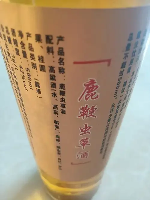 一瓶泡制好的琥珀红色蛹虫草鹿血酒特写，旁边摆放着干燥的蛹虫草和鹿茸片，背景为古朴的木桌和酿酒器具，整体氛围传统而健康。_1