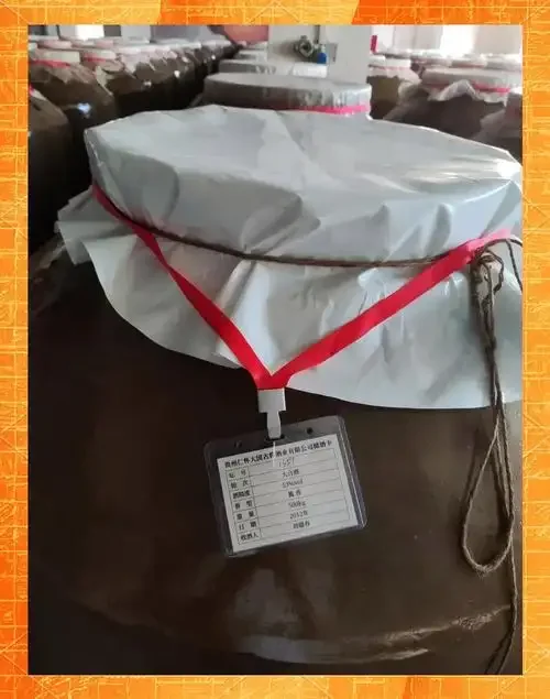 一个用红布封口的棕色陶瓷酒坛，旁边放着旧标签和工具，置于木质酒架前，展示了传统酱香白酒的储存场景。_1