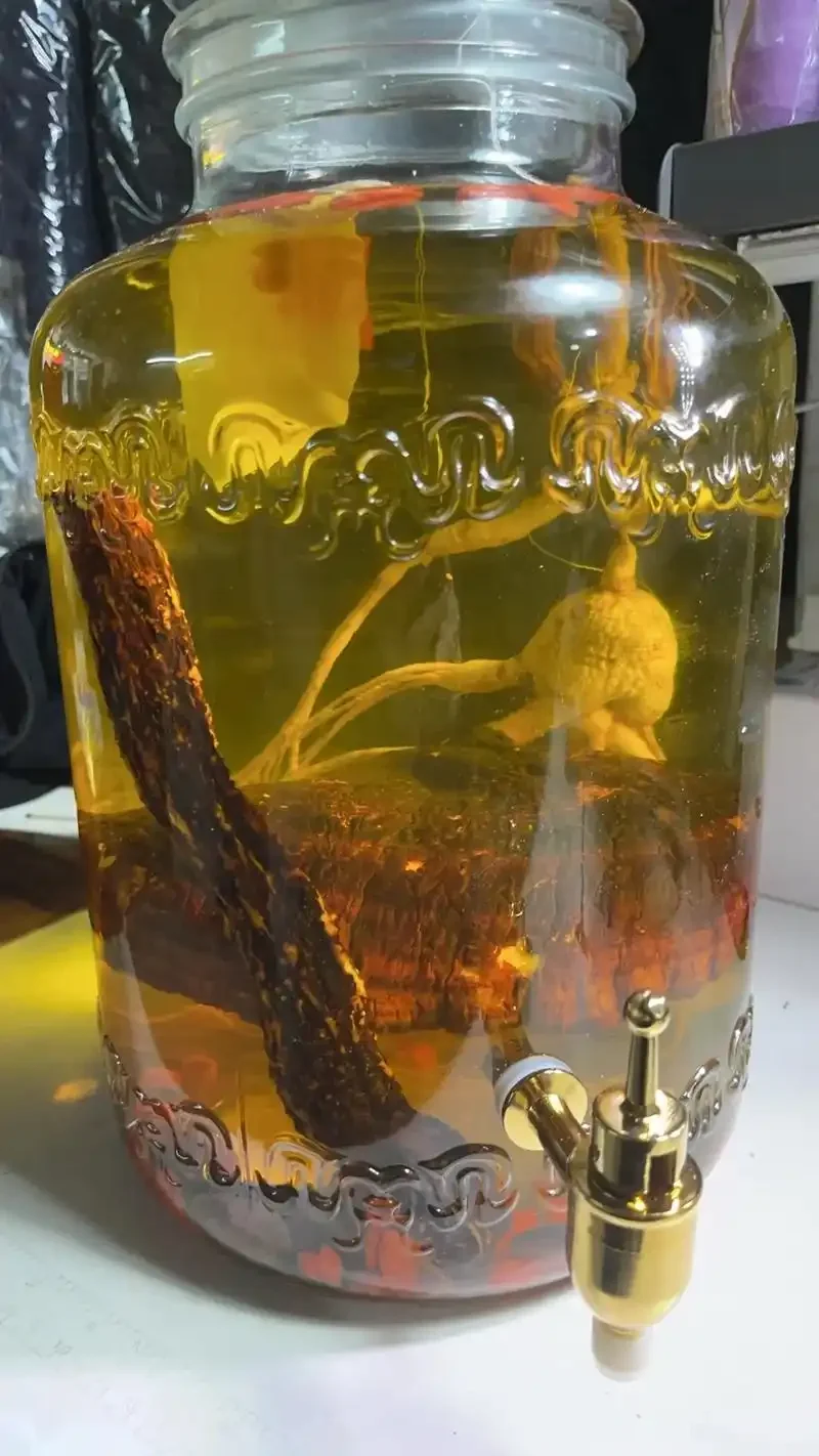 一个装有琥珀色酒液的透明玻璃罐，罐内清晰可见整根人参、切片灵芝和红色枸杞，旁边放着一小杯成品，展示家庭自制灵芝人参枸杞酒的过程。_1